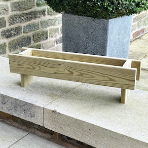 May include: A wooden planter box with a natural finish. The planter is made of unfinished wood and has a rectangular shape. The planter is sitting on a stone surface.