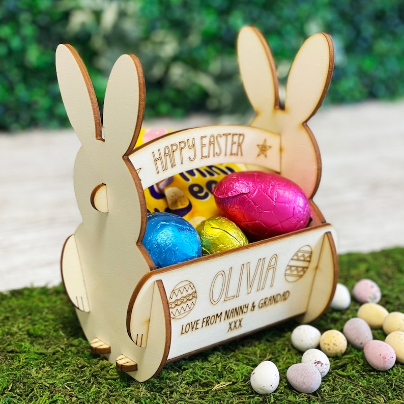 May include: A wooden Easter basket shaped like a bunny, filled with colorful foil-wrapped eggs and candy. The basket has the words "HAPPY EASTER" and "OLIVIA" engraved on it, along with the message "LOVE FROM NANNY & GRANDAD XXX".