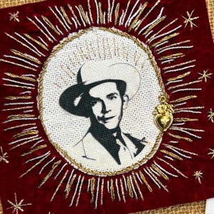 May include: A square dark red velvet patch featuring an embroidered portrait of a man in a cowboy hat surrounded by gold rays and a small gold heart charm. Beside it is a white rectangular card with the same portrait and text: HANK WILLIAMS for the faithful.