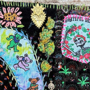 May include: A square, black fabric art piece with colorful embroidered designs. Features dancing bears, a Steal Your Face skull, and a skeleton with roses. Accented with gold charms and a tan border. The words "Grateful Dead" are visible.