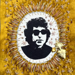 May include: A rectangular mustard yellow velvet textile piece featuring a central oval portrait of a person with dark hair wearing sunglasses. The portrait is surrounded by radiating metallic gold embroidery, small star-shaped accents, and a gold heart charm.