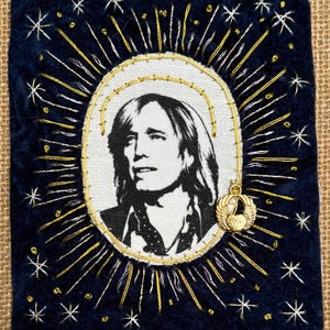 May include: A rectangular dark blue fabric patch featuring a central oval portrait of a person with shoulder-length hair. The portrait is surrounded by embroidered gold rays and white star motifs. A small gold winged heart charm is attached.