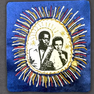 May include: A square blue fabric patch featuring a central cream-colored oval with a black and white illustration of two musicians, one playing a saxophone. The oval is surrounded by embroidered rays in red, white, and gold thread with a small heart charm.