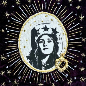May include: A square dark purple velvet textile piece featuring a central oval patch with a black and white portrait of a person wearing a star-patterned crown. The patch is surrounded by radiating gold and silver metallic embroidery and small stars.