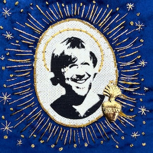 May include: A square blue fabric patch featuring a central oval portrait of a smiling person in black and white. The portrait is surrounded by gold embroidered rays and stars, with a small gold-toned heart charm attached to the lower right side.