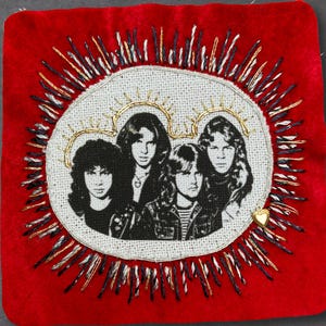 May include: A square red fabric patch featuring a central embroidered black and white portrait of a four-member rock band. The portrait is surrounded by a circular border of radiating black, white, and gold thread stitches and a small gold heart charm.