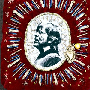 May include: A square red velvet textile piece featuring a central oval portrait of a person in profile, surrounded by embroidered gold and blue rays and stars. A small gold-toned metal heart charm and a yellow bead are attached to the right side.