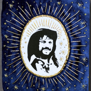 May include: A rectangular patch featuring a dark blue velvet background with gold embroidered sunburst rays and stars surrounding a central white oval. Inside the oval is a black and white portrait of a smiling person with a mustache and long hair.