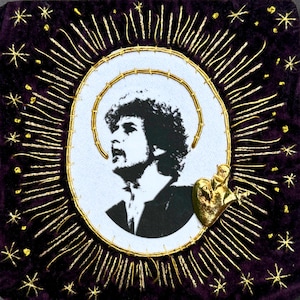 May include: A square purple velvet textile piece featuring a central white oval patch with a black and white portrait of a person with curly hair. The portrait is surrounded by gold embroidered rays, stars, and a gold metallic heart-shaped charm.