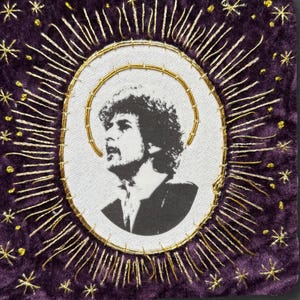 May include: A square purple velvet patch featuring a central white oval with a black and white portrait of a person with curly hair. The portrait is surrounded by gold embroidered rays and small gold stars on the purple fabric background.