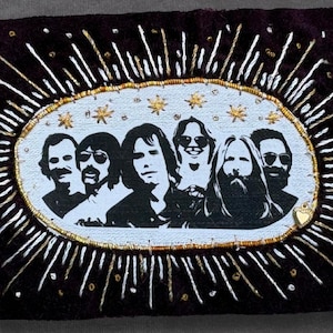 May include: A rectangular dark purple velvet patch featuring an embroidered oval center with a black and white illustration of six individuals. The oval is surrounded by radiating gold and white thread rays and small gold stars above the figures.