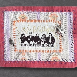 May include: A rectangular fabric patch featuring a central white panel with a black graphic of The Grateful Dead band members and text. The panel is bordered by orange stitching, a patterned floral fabric, and a red corduroy edge with purple stitching.