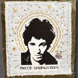 May include: A rectangular fabric patch featuring a black and white portrait of Bruce Springsteen on a white background. The design includes gold embroidered star accents, a gold halo, and the text BRUCE SPRINGSTEEN with a small metal faith charm.