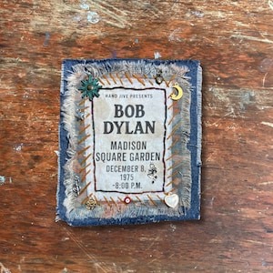 May include: A fabric patch with the text "BOB DYLAN MADISON SQUARE GARDEN DECEMBER 8, 1975 -8:00 P.M." is surrounded by a frayed blue border. The patch is embellished with small charms and decorative stitching.