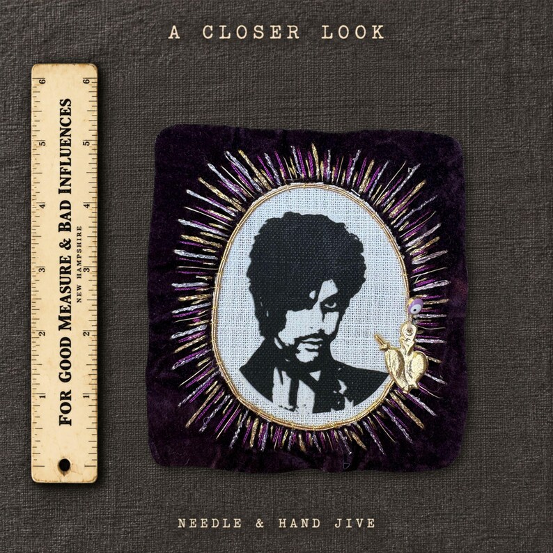 May include: A square dark purple velvet patch featuring a black and white embroidered portrait of Prince surrounded by radiating purple and gold metallic threads. A small gold heart charm is attached. A ruler shows a height of 5 inches.