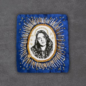May include: A square, blue fabric patch with an embroidered portrait of a person with long hair, surrounded by a gold halo and sunburst design. The portrait is in black and white, with gold and white accents.