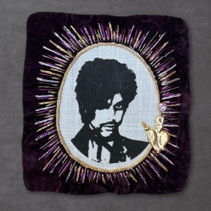 May include: A square dark purple velvet textile piece featuring a central oval embroidery of a person with curly hair in black thread. The oval is surrounded by radiating gold and purple metallic threads and a small gold heart charm with an eye bead.