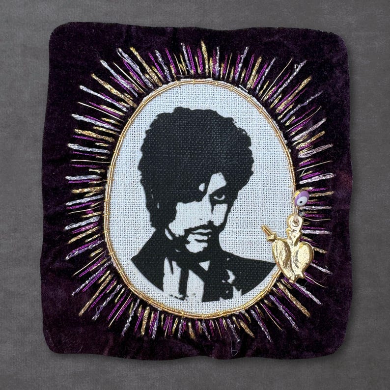 May include: A square dark purple velvet textile piece featuring a central oval embroidery of a person with curly hair in black thread. The oval is surrounded by radiating gold and purple metallic threads and a small gold heart charm with an eye bead.