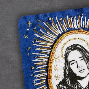 May include: Embroidered patch with a portrait and halo. The design features gold and white thread, a blue border, gold crosses, and star-like accents. The artwork has a religious or iconographic theme, with a detailed and ornate appearance.