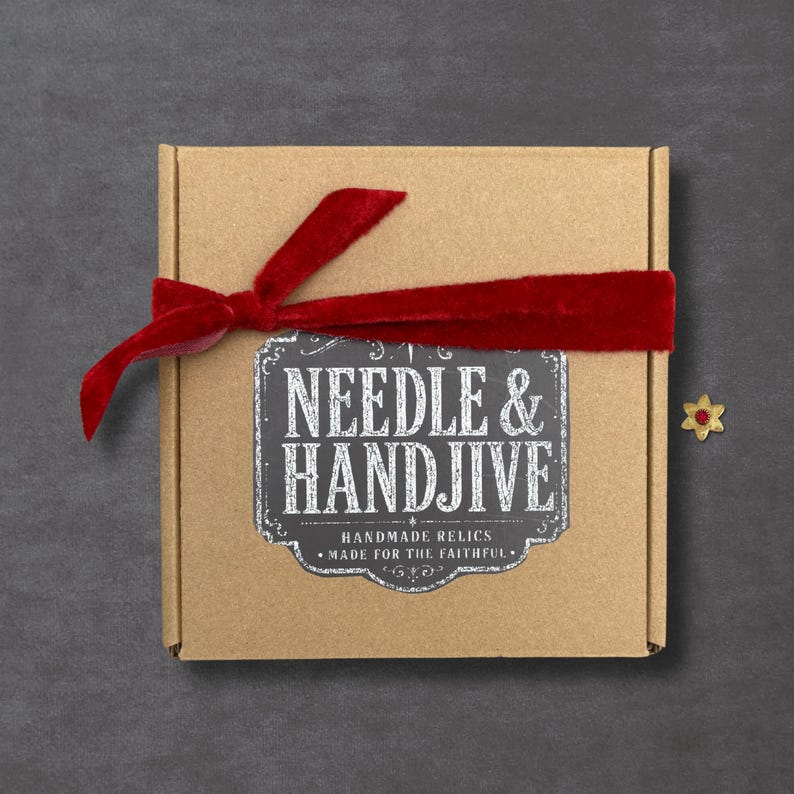 May include: A square brown cardboard box tied with a red velvet ribbon. A black label on the center features the white text "NEEDLE & HANDJIVE" above "HANDMADE RELICS" and "MADE FOR THE FAITHFUL." A small gold star-shaped pin with a red center sits nearby.