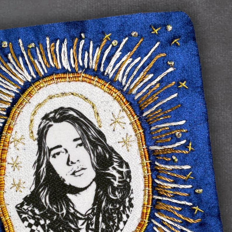May include: A blue embroidered patch with a black and white portrait of a person with long hair, encircled by a gold halo and radiating lines. The design incorporates gold and white thread and star-shaped embellishments.