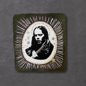 May include: A square, olive green patch with a black and white image of a person playing a guitar. The image is surrounded by a white oval with a halo of silver and gold thread. The patch is likely an embroidered item.