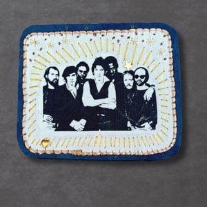 May include: Embroidered patch featuring a black and white image of a group of men. The patch has a blue border with gold stitching and a light background with gold starburst and star accents. A gold heart is at the bottom.