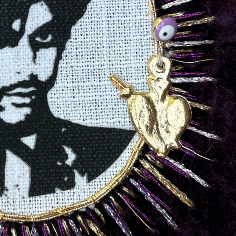 May include: A textile art piece featuring a black and white portrait of a person on a circular fabric patch. The patch is bordered by metallic gold and purple fringe, accented with a small gold heart charm and a tiny purple evil eye bead.