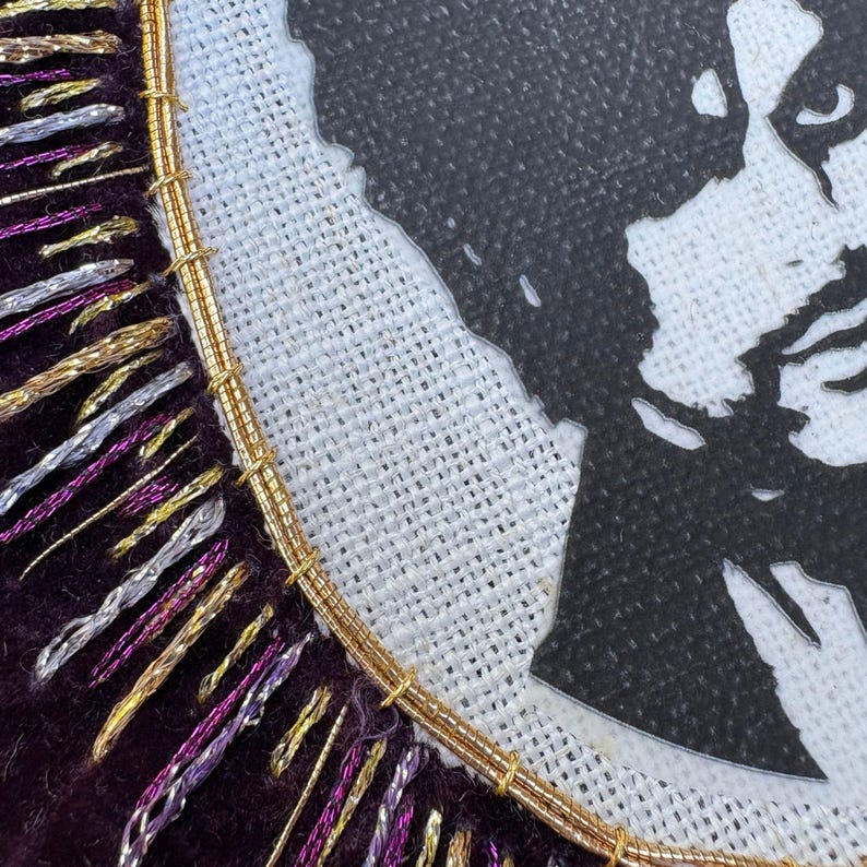 May include: A circular embroidery piece featuring a black silhouette of a person on a white textured fabric. The border is made of dark purple velvet fabric adorned with radiating metallic gold, silver, and purple threads stitched in a fringe pattern.