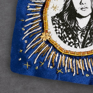 May include: A close-up of a blue velvet embroidered patch featuring a black and white portrait. The patch is decorated with gold, white, and silver thread, and a small gold flower. The portrait is framed by a gold and brown beaded border.