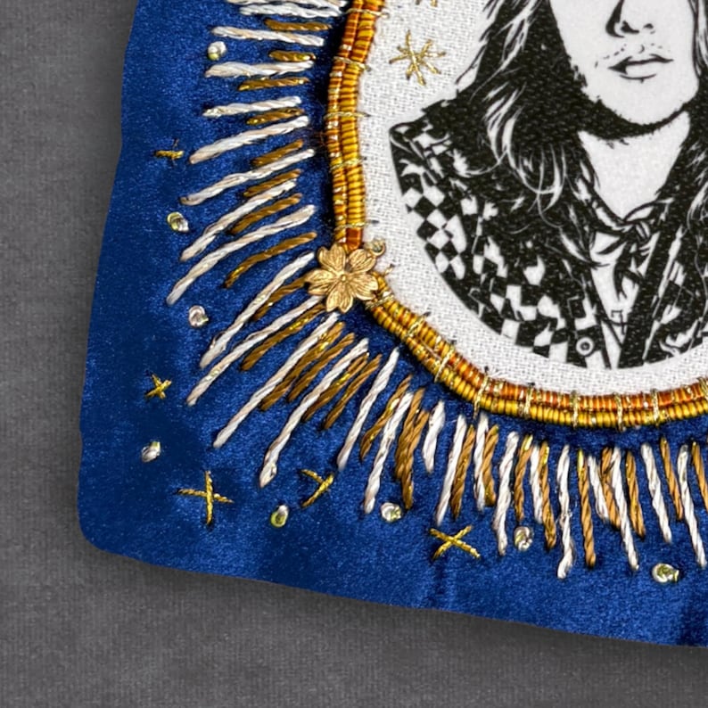 May include: A close-up of a blue velvet embroidered patch featuring a black and white portrait. The patch is decorated with gold, white, and silver thread, and a small gold flower. The portrait is framed by a gold and brown beaded border.