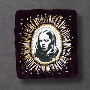 May include: A dark burgundy velvet patch featuring a black and white portrait within an oval frame. The frame is surrounded by gold and white embroidered rays and small, scattered white beads. A small gold heart is at the bottom.
