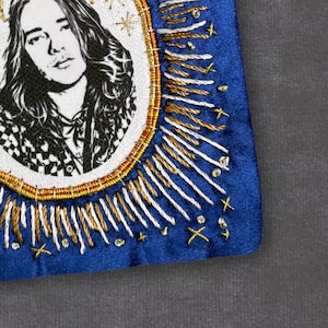 May include: Embroidered portrait of a person with long hair, in a black and white checkered shirt, within an oval frame. The frame is surrounded by gold and white stitching on a blue background, with star and cross accents.