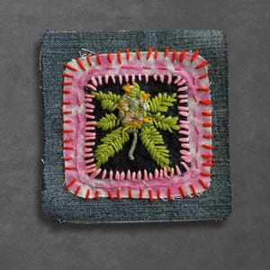 May include: Embroidered patch featuring a cannabis leaf design. The patch is square, with a denim base and a black center. The leaf is green and yellow, surrounded by a pink border and red stitching. A unique, handmade accessory.
