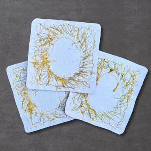 May include: Three square white fabric coasters featuring a central circular design embroidered with metallic gold thread. The gold stitching creates a textured, abstract pattern that radiates outward from the center of each square piece.