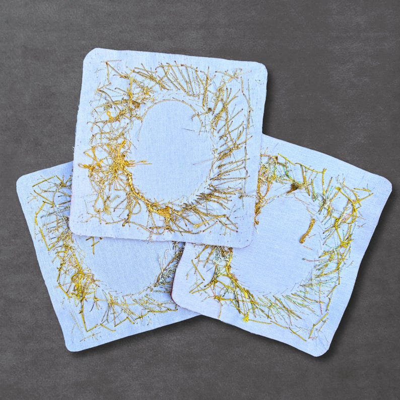 May include: Three square white fabric coasters featuring a central circular design embroidered with metallic gold thread. The gold stitching creates a textured, abstract pattern that radiates outward from the center of each square piece.