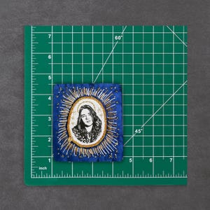 May include: A blue fabric patch with a black and white portrait inside a gold halo design. The patch is approximately 7.6 cm tall and 6.3 cm wide. Gold and white thread embroidery surrounds the portrait and the patch's edges.