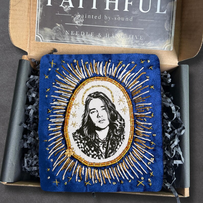 May include: A blue embroidered artwork with a portrait, gold halo, and sunburst design. The piece is presented in a box with the text "FAITHFUL" and "NEEDLE & HAND JIVE". Decorative packing material surrounds the artwork.