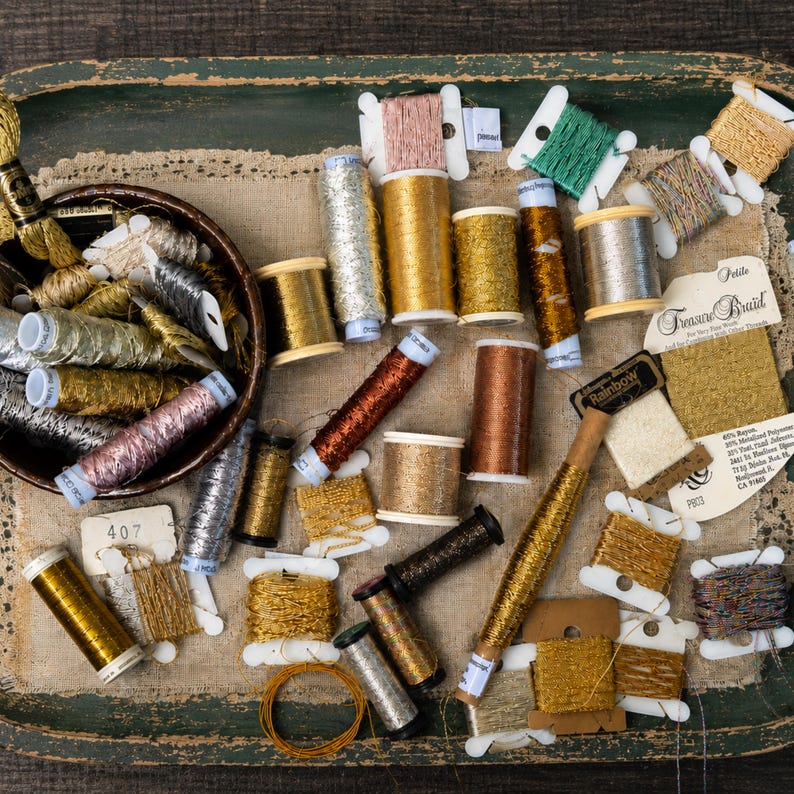 May include: A collection of metallic embroidery threads in various shades of gold, silver, copper, and bronze. The threads are presented on spools, bobbins, and cards, with some contained in a dark bowl. One card is labeled Treasure Braid.