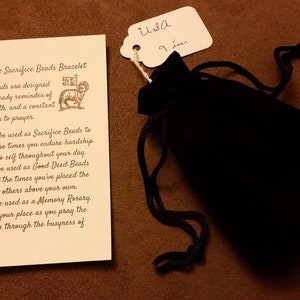 Puede incluir: Una tarjeta blanca con texto marrón que describe un brazalete de cuentas de sacrificio simple. El texto dice "Simple Sacrifice Beads Bracelet. Estas cuentas están diseñadas para ser un recordatorio constante de tu fe y una invitación constante a la oración. Se pueden usar como cuentas de sacrificio para contar las veces que soportas dificultades o mueres a ti mismo a lo largo de tu día. Se pueden usar como cuentas de buenas acciones para contar las veces que has puesto las necesidades de los demás por encima de las tuyas. Se pueden usar como un rosario de memoria, manteniendo tu lugar mientras rezas los misterios en medio del ajetreo de la vida diaria." Una pequeña bolsa de terciopelo negro está al lado de la tarjeta con una etiqueta blanca que dice "USA 9 ine".