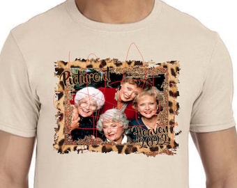 May include: A beige t-shirt featuring a graphic print of four women from the television show "Golden Girls." The graphic is surrounded by a leopard print frame and the text "Picture it... Heaven 2022."