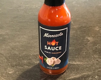 Może przedstawiać: Butelka sosu ostrego Minnesota o pojemności 148 ml z czarną etykietą i białym obrazkiem czosnku. Etykieta zawiera napis "Minnesota Hot Sauce Garlic Habanero".