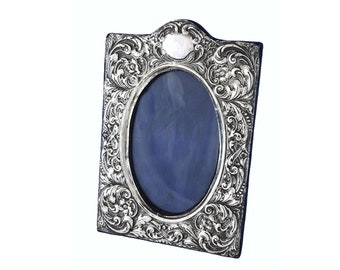 May include: An ornate silver picture frame with a detailed floral design. The frame has an oval opening and a raised, circular design at the top.