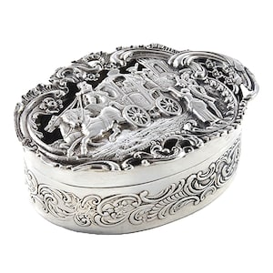 May include: An oval silver-coloured jewellery box with a detailed relief scene of a horse-drawn carriage. The box features ornate scrollwork around the edges and on the lid, creating a vintage aesthetic. The lid's design includes a carriage, horses, and figures.