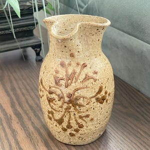 May include: A beige ceramic pitcher with a speckled texture and a brown hand-painted floral design on the front. The pitcher features a rounded body, a flared rim, and a small pouring spout, showcasing a natural, earthy aesthetic throughout.