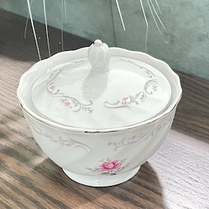 Op de afbeelding: Een witte porseleinen suikerpot met bijpassend deksel, voorzien van een delicaat roze rozenpatroon en grijze krulmotieven. De kom heeft een geschulpte rand met een zilverkleurige metalen afwerking en een kleine, ronde knop op het deksel.