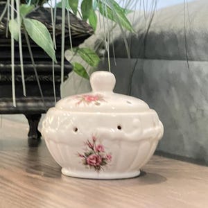 May include: A small, round, cream-colored ceramic bowl with a matching lid. The bowl features a delicate floral pattern of pink roses and green leaves, with a series of small, evenly spaced circular ventilation holes around the upper rim.