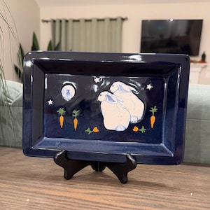 May include: A rectangular dark blue ceramic tray featuring a raised design of two white rabbits, orange carrots, small green leaves, and white stars against a night sky background, displayed on a black plastic stand.