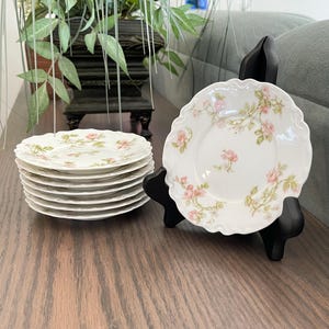 May include: A set of seven white porcelain saucers featuring a delicate pink rose and green leaf floral pattern. Each saucer has a scalloped edge. One saucer is displayed upright on a black plastic stand, while six are stacked in a pile.