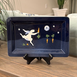 May include: A rectangular dark blue ceramic tray featuring a white rabbit leaping through a night sky filled with stars and a full moon. The rabbit holds an orange carrot, while two additional carrots are depicted below. The tray rests on a black stand.